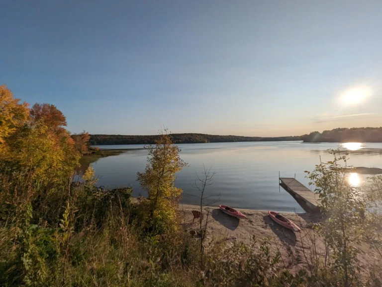 Autumn leaves on the trees framing a quiet lake, with soft reflections on the water at sunset. 2 Kayaks rest beside the dock.