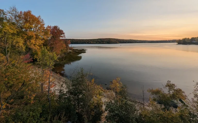A calm lake framed by trees with the sun low on the horizon and two kayaks on the shore.
